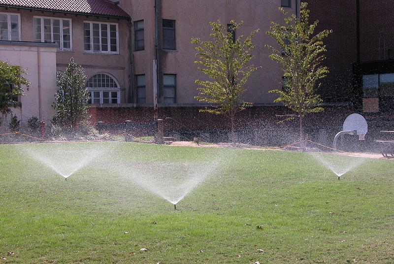 三个洒水器正在给草坪浇水，背景是建筑物和树木