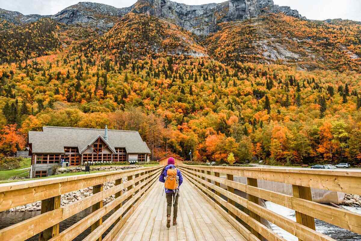 背包客走过一座桥，走近山谷里的一个农场，背景是覆盖着五彩秋叶的小山