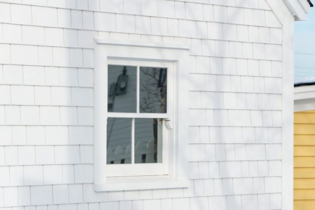 一座老式木制建筑的外角，有一扇封闭的玻璃单挂窗。墙上覆盖着白色的瓦片。背景中有一座白色镶边的黄色木制建筑。